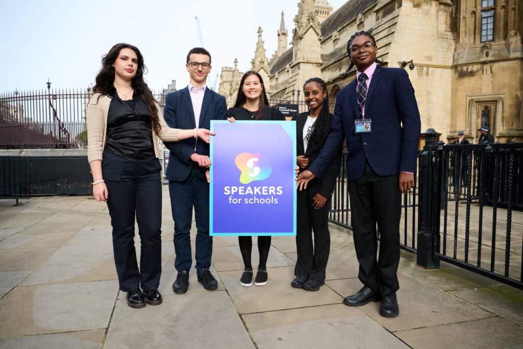 A group of five young people, dressed in business attire, stand outside London's House of Commons holding a large "Speakers for Schools" sign.